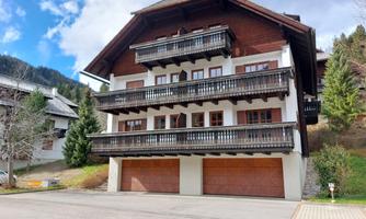 A cozy chalet in alpine style with balconies and wooden cladding. In the foreground, there is an asphalt road and some greenery.