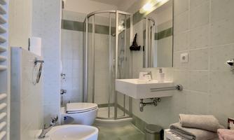 A modern bathroom with a shower, a sink, and a toilet. The walls are painted in soft green tones, and there is a shelf with towels.