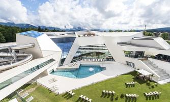 A modern building with a swimming area and a slide.  
Surrounded by a large green space with sun loungers and a clear sky.