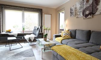 A modern living room with a gray sofa and colorful decorative cushions. There are large windows that let in plenty of light, as well as decorative plants and pictures on the wall.
