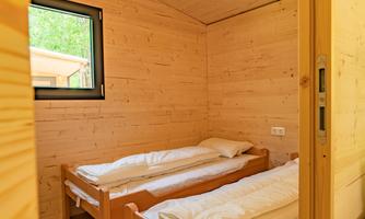 A cozy room with two wooden single beds. The walls are made of light wood, and a window lets in daylight.