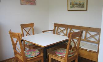 A cozy dining area with a white table and wooden chairs. In the background, there is a picture and a bench.