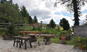 A cozy outdoor area with wooden tables and benches. In the background, there is a green landscape with trees and a blue sky.