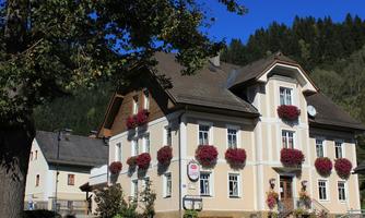 A picturesque building with colorful flower boxes at the windows. In the background, green mountains and a blue sky are visible.