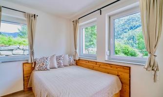 A bright bedroom with a large bed and colorful pillows. The windows offer a view of the green landscape outside.