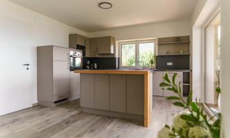 A modern kitchen with wooden and gray elements. It features a kitchen island and large windows that let in plenty of light.