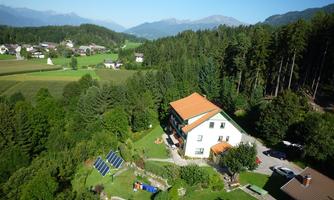 A picturesque house with a red roof, surrounded by green spaces and trees. In the background, mountains and a quiet village can be seen.