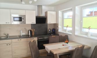 A modern kitchen with wooden cabinets and a bright dining area. The table is decorated with a bouquet of flowers and there are large windows that let in plenty of light.