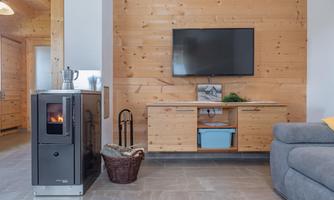 A cozy living room with a wood stove, a television on the wall, and a gray sofa. The walls are made of wood and give the room a warm atmosphere.