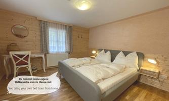A cozy bedroom with a large bed and wood paneling. A table and chair complement the room, while daylight streams through the window.