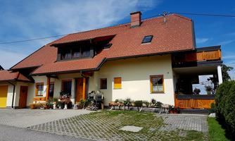 A large, two-story house with a red roof and yellow windows. In front of the house, there are flowers and a paved area.