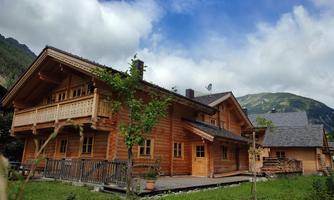 A charming wooden house surrounded by natural vegetation and mountains. The sky is partly cloudy, creating a relaxed atmosphere.