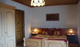 A cozy bedroom with a double bed and typically rustic wooden furniture. The walls are painted light, and a decorative lamp provides warm light.