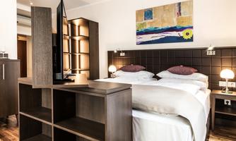 A modern hotel room with two single beds and a flat-screen TV. The walls are decorated with a colorful piece of artwork, and there is warm light from the lamps.