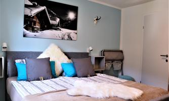 A cozy bedroom with a large bed and colorful pillows. On the wall hangs a picture of a mountain cabin in the snow.