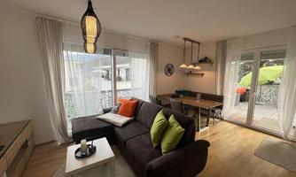A cozy living room with a dark gray sofa and colorful cushions. Large windows offer a view of a terrace with green furniture.