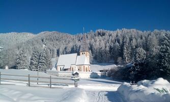 A snow-covered landscape with a small, picturesque village and a church. The clear blue sky and the snow-covered trees create a wintry atmosphere.