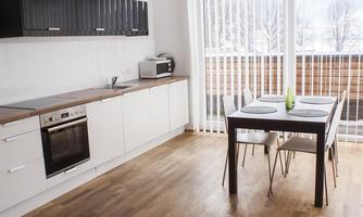 A modern kitchen with white cabinets and wooden shelves. A dining table with four chairs is situated near a large window with curtains.