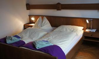 A cozy bedroom with two beds, each with a white blanket and a purple bed sheet. Next to the beds are nightstand lamps and a small table.