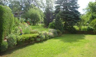 A well-maintained garden with green grass, colorful flowers, and large trees. In the background, more plants and bushes are visible.