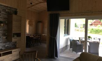 A cozy living space with wood paneling and modern furniture. In the background, there is a dining area and large windows with a view outside.