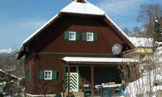 A cozy wooden house in winter, surrounded by snow. The windows are painted green and the roof is snow-covered.