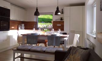 A modern kitchen with bright wood and elegant lighting. The dining area is inviting and well-lit by windows.