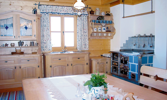 A cozy kitchen with wooden cabinets and a large dining table. Bright windows and a traditional stove create an inviting atmosphere.