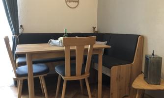 A cozy dining area with a wooden table and chairs. The bench is designed in dark fabric, and a window with curtains allows for daylight.