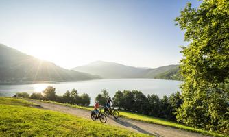Two cyclists ride along a path by the lake. The sun is shining and the mountains are visible in the background.