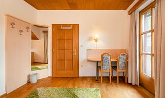 A simple room with wooden flooring and light walls. There is a desk, two chairs, and a large window with curtains.