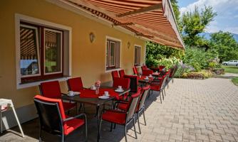 A cozy terrace with red chairs and tables. The surroundings are green and inviting, ideal for relaxed hours outdoors.