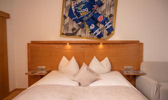 A modern bedroom with a wooden bed and three pillows. A striking picture of a blue tracksuit hangs on the wall.