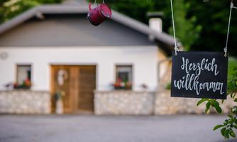 An inviting house with a welcome sign in handwritten script. In the foreground, a small red kettle hangs from a tree.