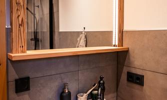 A modern bathroom with a round ceramic sink and wood details. The mirror is illuminated with warm light, and the wall is in neutral tones.