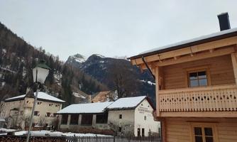 An idyllic mountain landscape with snow-covered roofs and wood piles. In the background, there are tall mountains and a cloudy sky.