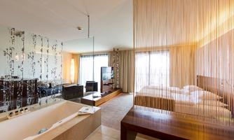 A modern hotel room with a cozy bed and a separate living area. The large windows provide plenty of natural light.