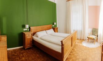 A cozy bedroom with a double bed and a green accent wall. Bright furniture and a chair complement the relaxed atmosphere.