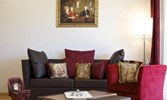 A cozy living room with a dark-toned sofa and red cushions. A classic painting hangs on the wall, and a lamp stands next to the sofa.
