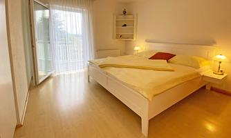 A bright bedroom with a large bed and two nightstands. The windows let in a lot of daylight and lead to a small balcony.