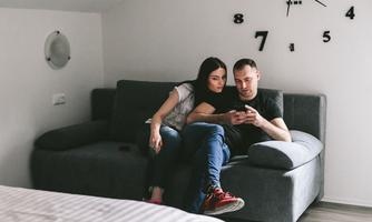 A couple is sitting on a gray sofa, looking at a mobile phone together. The living environment is modern and bright.