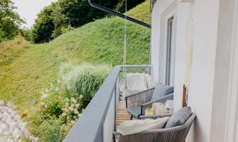 A balcony with two comfortable chairs and a table. In the background, green hills and plants can be seen.