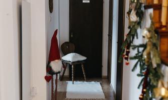 A bright hallway with wooden floors and white carpets. Christmas decorations hang on the wall, and a chair stands next to a door.