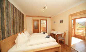 A cozy room with two beds, wooden decor, and large windows. There is a small table with chairs and access to a balcony.