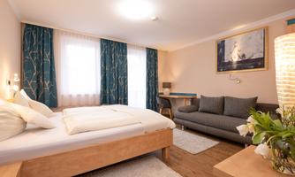 A modern hotel room with a double bed and a cozy seating area. The walls are light, and there are large windows with blue curtains.