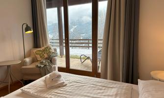 A cozy bedroom with two beds and a balcony. The view shows a mountainous landscape and gray clouds.