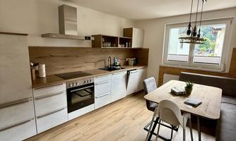 A modern kitchen with wooden shelves and white units. A dining table with chairs accommodates several people.