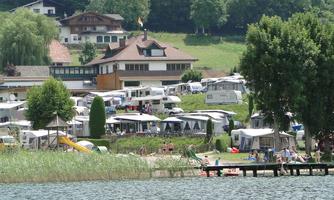 A campsite by the lake with caravans and a beautiful house. In the foreground, people are sitting by the shore and enjoying nature.