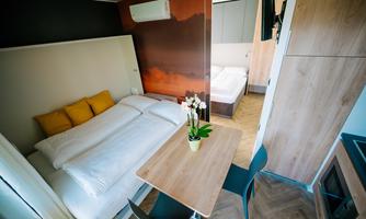 A modern bedroom with a double bed and a small table. In the background, a second bed can be seen, and there is a bright, pleasant atmosphere.