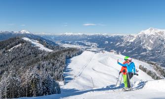 Two skiers stand on a snow-covered mountainside and enjoy the view. In the background, impressive mountains and a clear blue sky can be seen.